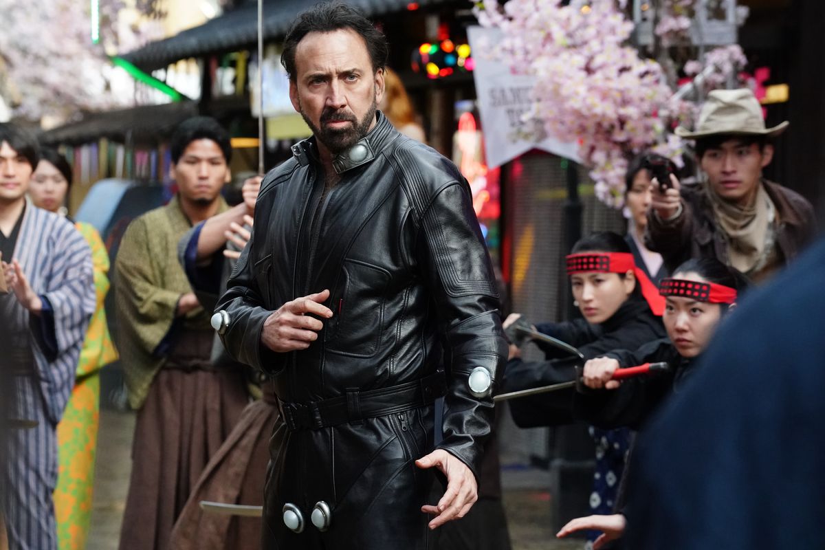 Nicolas Cage in a leather suit preparing to karate chop someone as a crowd watches in Prisoners of the Ghostland