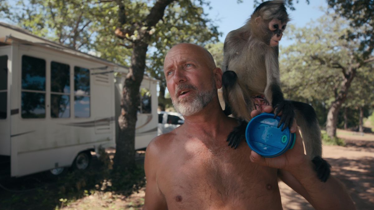 Wildlife owner Tim Stark, shirtless and with a monkey perched on his shoulder, standing in front of a trailer in Tiger King 2