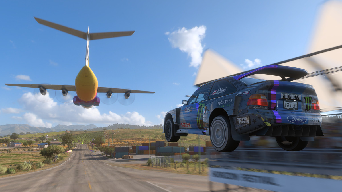 A rally car launches off a ramp with a large cargo plane in the distance