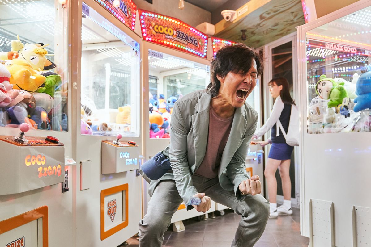 A man in a gray jacket and pants hunches over screaming in an arcade in Squid Game