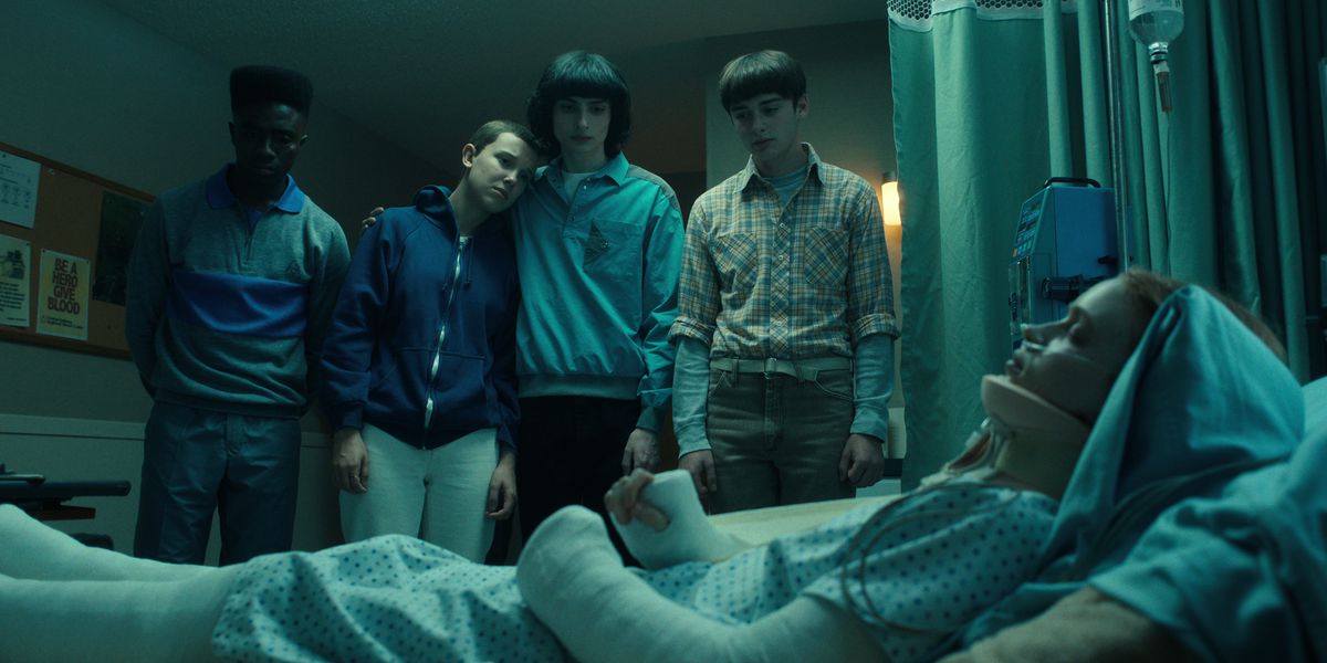 Lucas, Eleven, Mike, and Will standing and looking at Max in a hospital bed