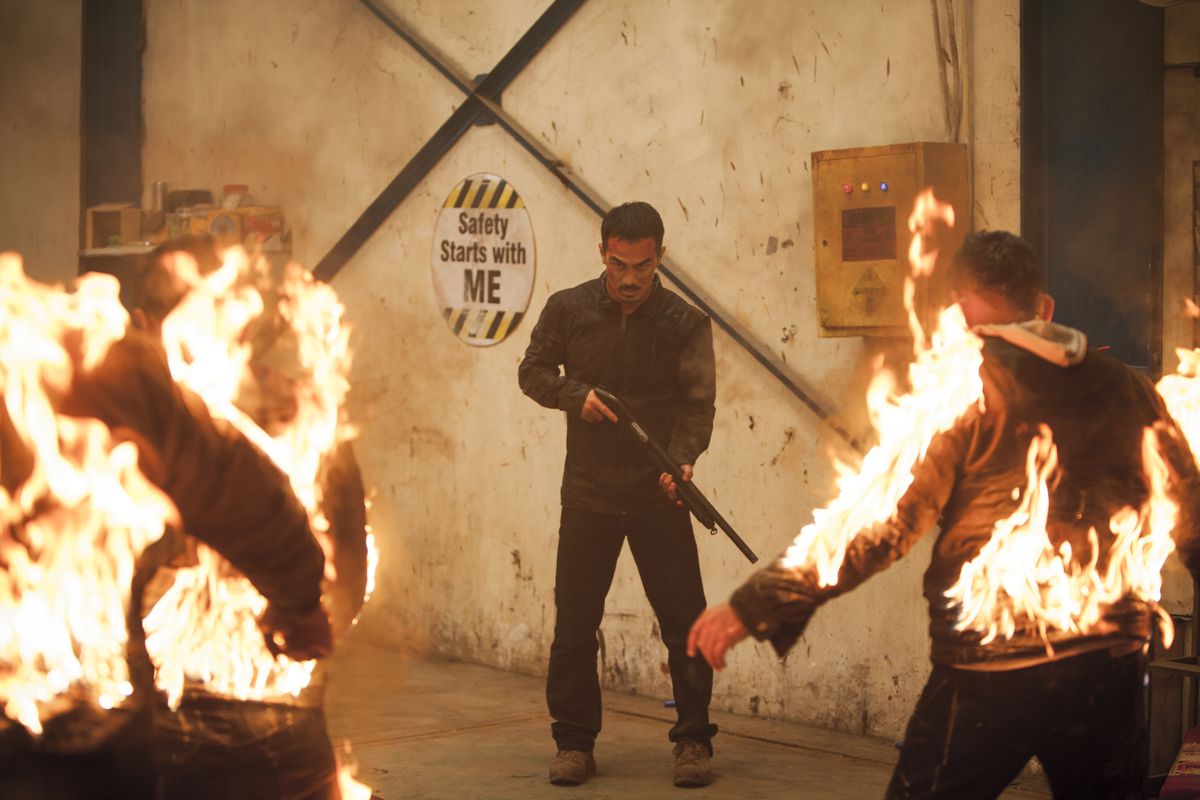 Joe Taslim stands in front of a “Safety starts with me” sign touting a shotgun facing several men on fire in The Night Comes for Us.