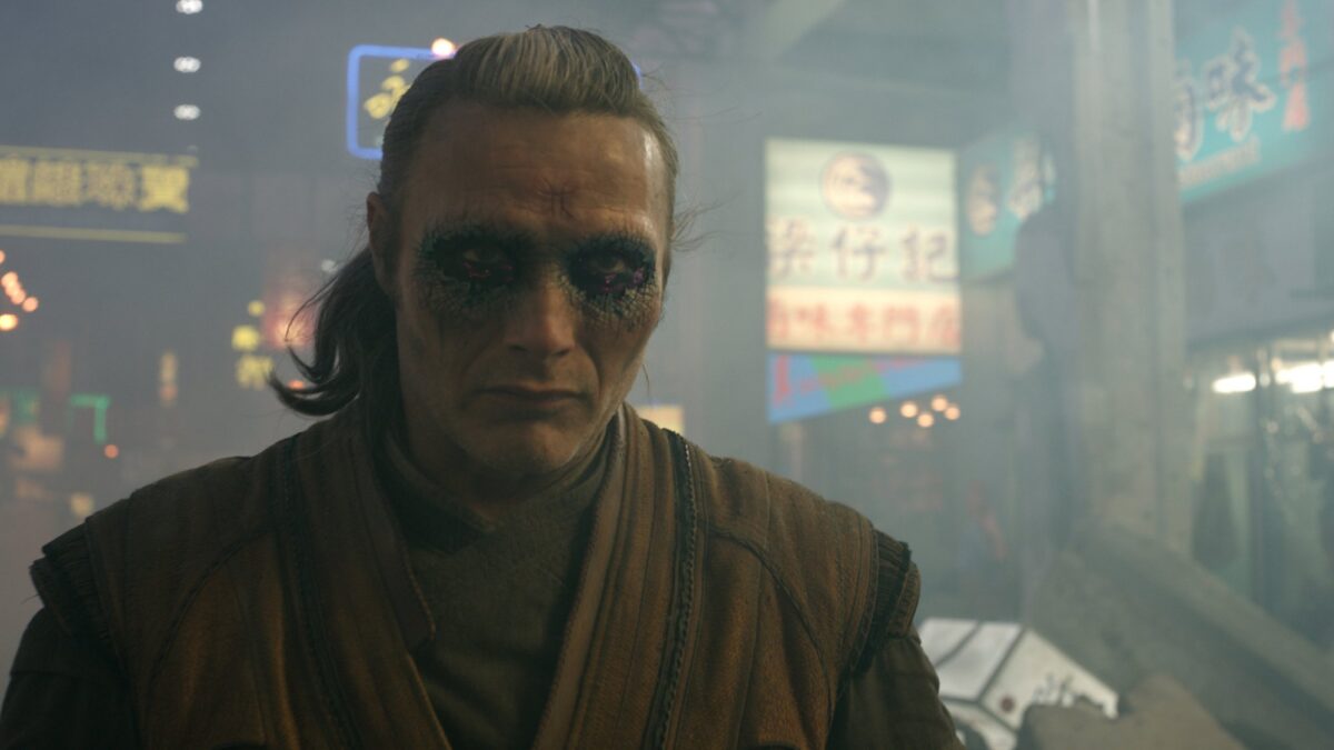 A man with dark makeup around his eyes and a low, short ponytail, wearing robes. In the background, the city of Hong Kong under attack.