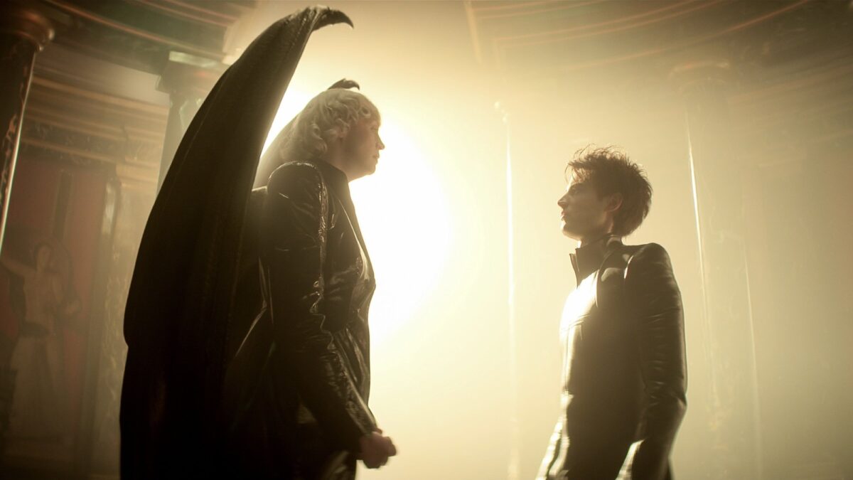 A winged woman in black and a man face each other in front of a blinding gold light.