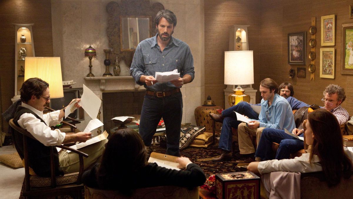 Ben Affleck as Tony Mendez standing in a room of people with a script in his hand in Argo.