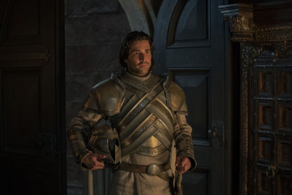 Ser Criston Cole stands at the entrance of a room wearing his knight armor and holding his helmet in House of the Dragon.