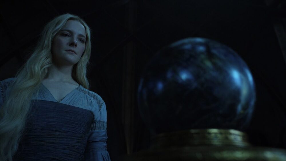 Galadriel, a blonde elf in a blue dress, stares at a glass orb on a gold pedestal.