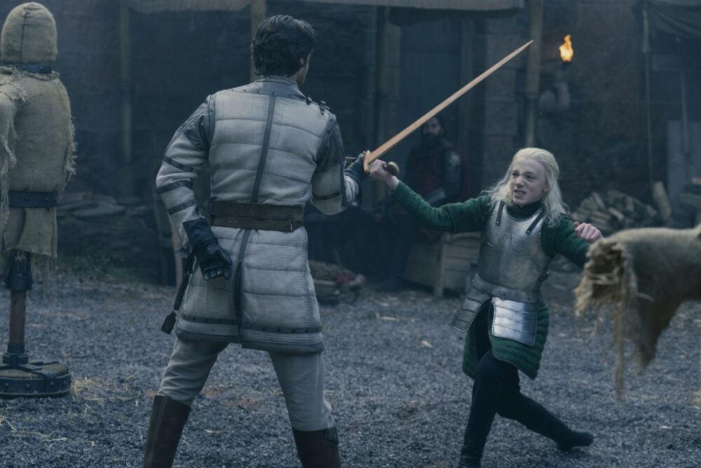 Leo Ashton as Aemond Targaryen, losing a sword fight. He has long bright hair and wears a green outfit covered by plate armor.