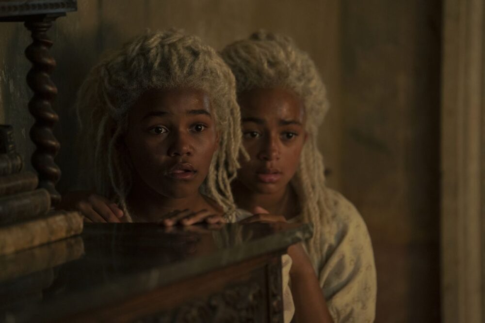 Shani Smethhurst and Eva Ossei-Gerning from HBO’s House of the Dragon as Baela Targaryen and Rhaena Targaryen. They have bright locs, and peer out from behind a dresser.