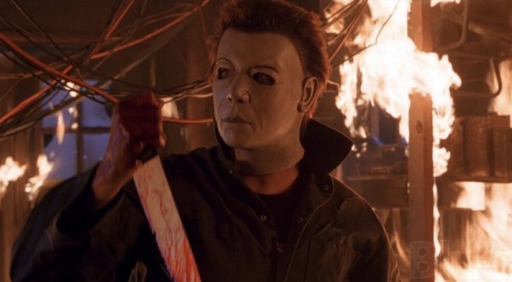 Michael Myers, surrounded by flames, wears a mask with arched eyebrows and a more distinct nose. He holds a bloody knife.