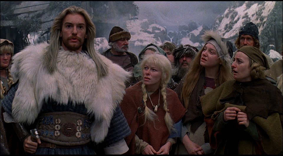 A group of Viking villagers stand amongst snowy mountains, with one man standing tall with white fur and long blonde hair, in Erik the Viking.