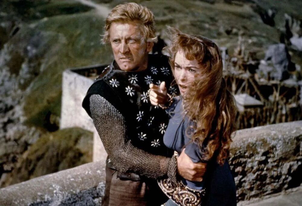 A man in viking clothing with a blind eye (Kirk Douglas) clutches the eyes of a shouting woman overlooking a stone balcony.