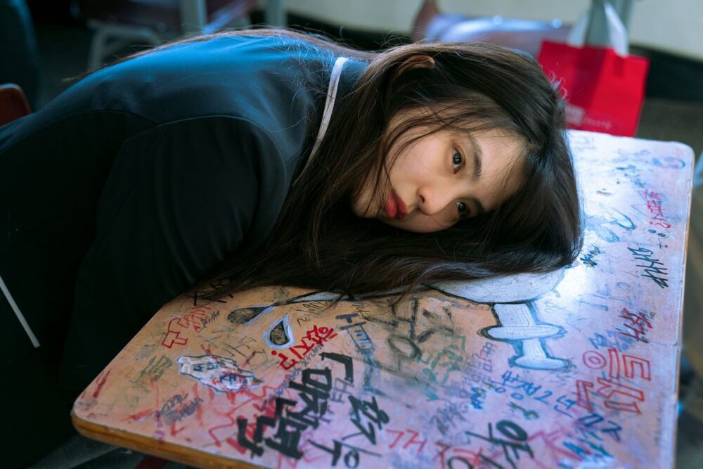 Han So-Hee rests her head against a desk with lots of drawings on it in My Name.