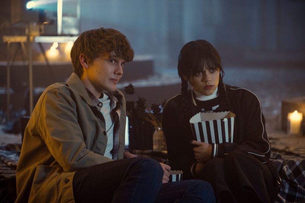 hunter, a pale young man with scruffy hair, sits next to wednesday, a young woman with dark hair in braids. she holds popcorn. they appear to be in some sort of crypt