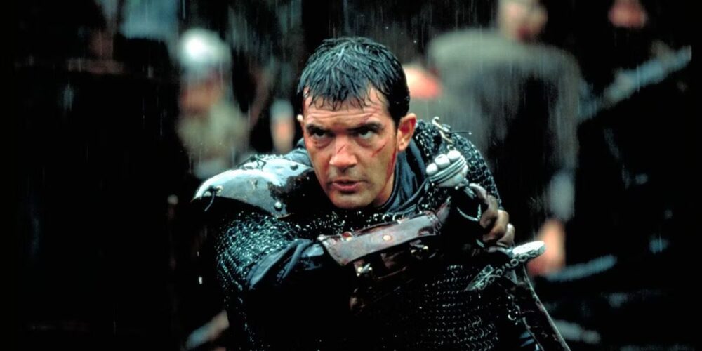 A man with visible scars on his face (Antonio Banderas) draws his sword from its sheath while drenched in rain.