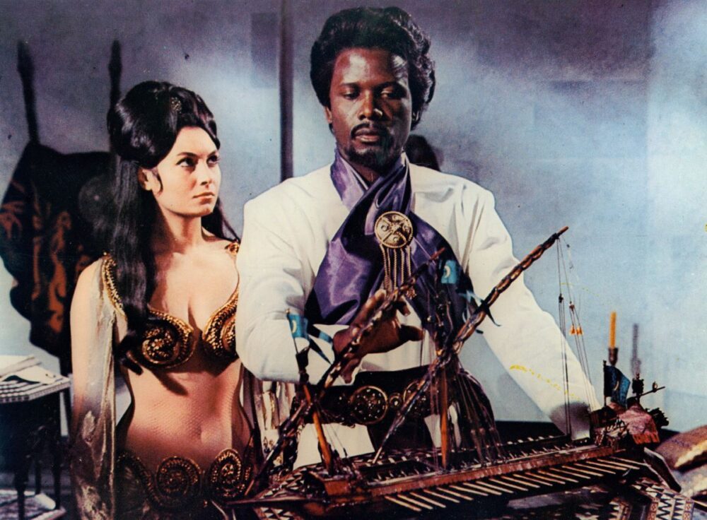 Sidney Poitier in fancy garb looking at a model ship next to a woman in The Long Ships.