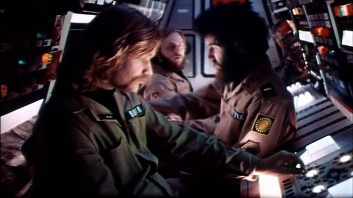 Three men in matching uniforms sit back to back against one another in a cramped spaceship cockpit.