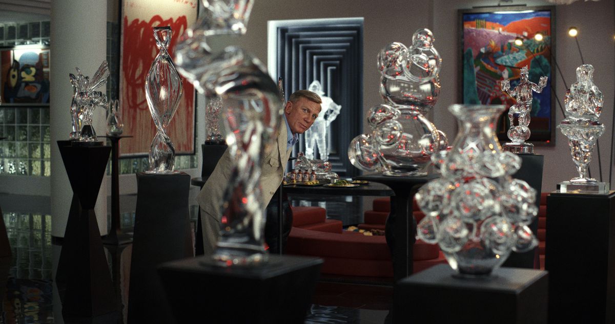 Detective Benoit Blanc (Daniel Craig) peers through a series of glass sculptures on ricky-looking pedestals in an art-packed room in Glass Onion