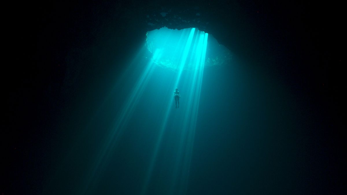 a deep dark ocean scene illuminated by a beam of turquoise water as light streams through a cave opening. one small diver swims in the middle