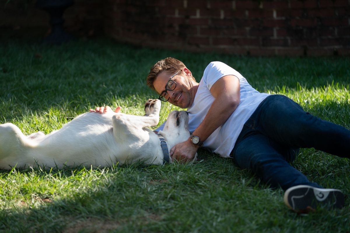 A man (Rob Lowe) wearing glasses, a white t-sthirt and blue jeans laying in a patch of grass and playing with a dog.