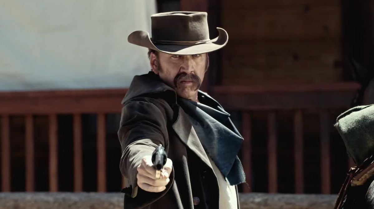 A man with an overgrown moustache (Nicolas Cage) wearing a cowboy hat and dark brown duster coat aiming a pistol.