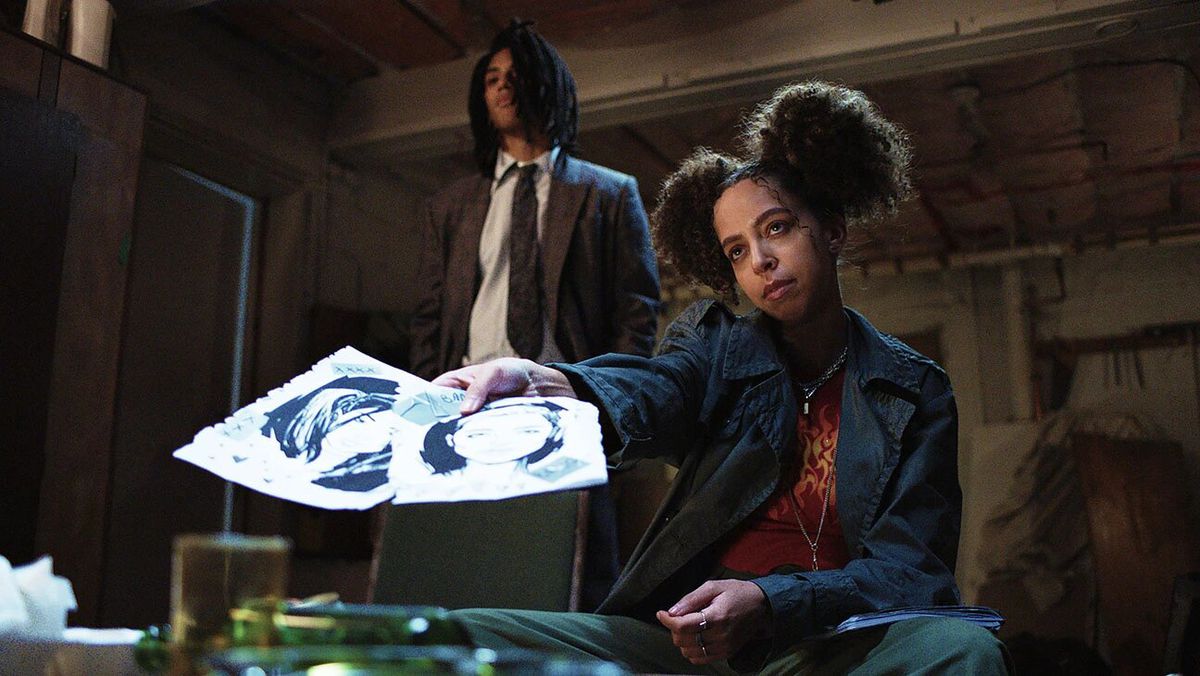 A woman with hair poofs (Hayley Law) holds out two drawings while seated behind a table.