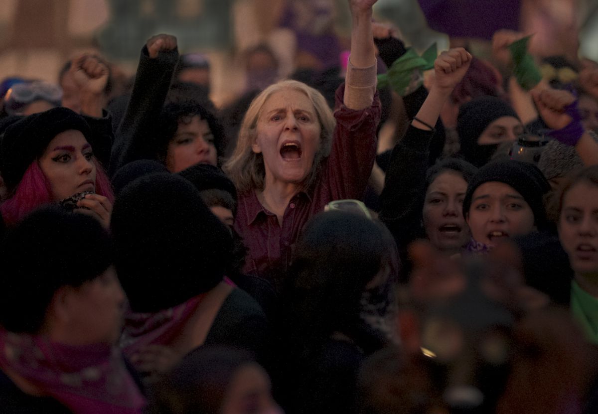 An older woman with gray hair shouting and raising her fist in a crowd of angry women.