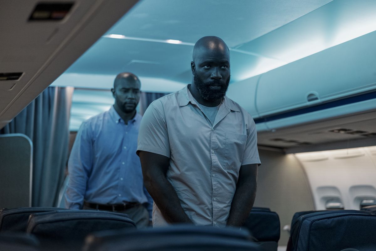 Louis Gaspare (a bearded Mike Colter) walks in handcuffs down the middle aisle of a plane with his cop escort following behind in Plane