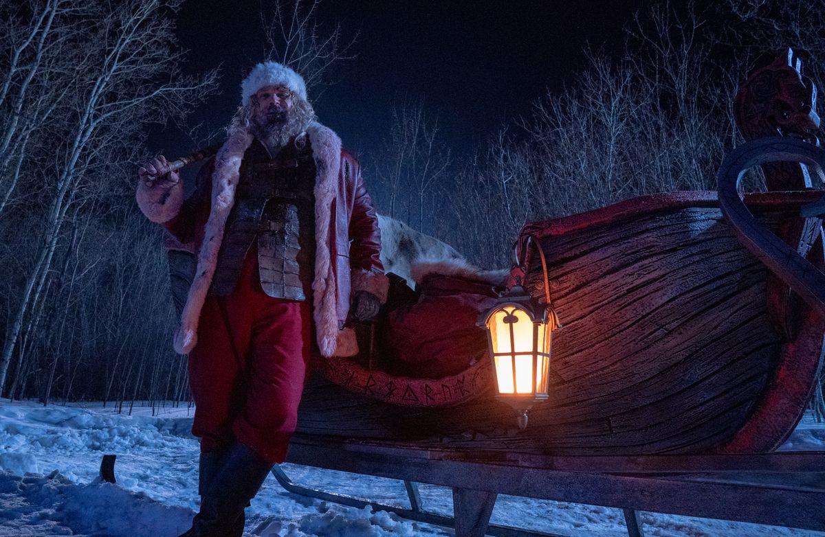 Santa Claus (David Harbour) leans drunkenly against his sleigh, a blood-red wooden boat-shaped vehicle carved with Nordic runes, in Violent Night