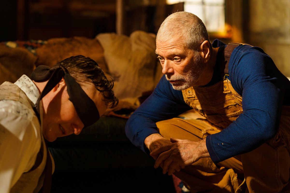 An old man (Stephen Lang) in a long-sleeve blue shirt and yellow overalls squats in front of a blind-folded man (Marc Senter).