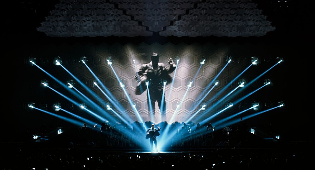 Justin Timberlake performs on stage, and a larger version of him appears on the screen behind him, blanketed by light blue lights, in Justin Timberlake + The Tennessee Kids.