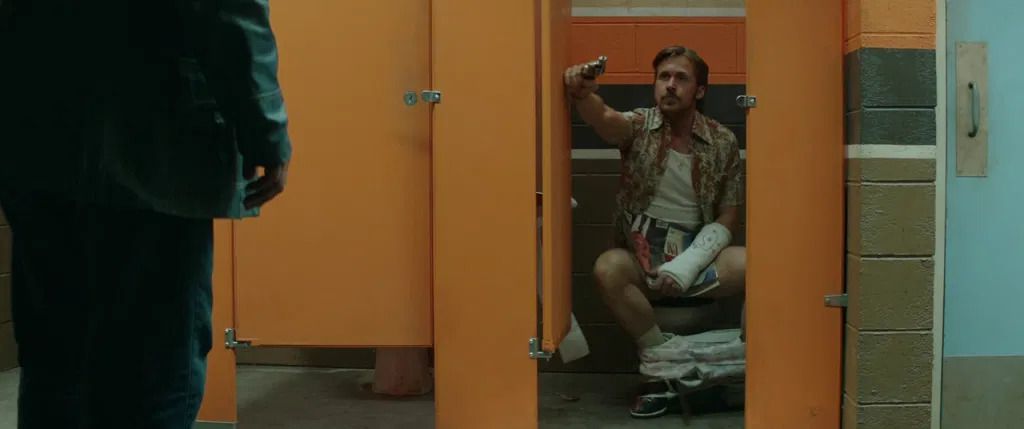 A man sitting in a orange bathroom stall with his pants down holding a door open with a pistol in one hand and a cast on his left arm stares at a man wearing a blue jacket and jeans to his right.