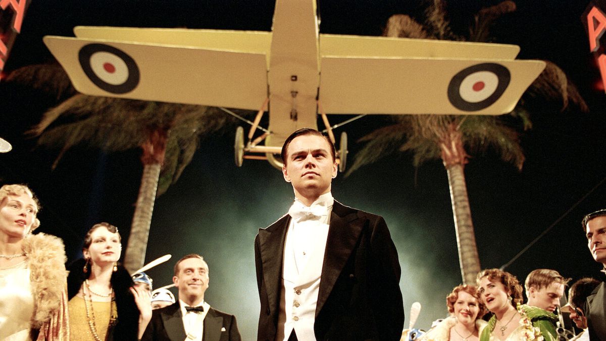 A man in a tuxedo (Leonardo DiCaprio) stands beneath a yellow biplane suspended above him while flanked by rows of extravagantly dressed onlookers and palm trees.