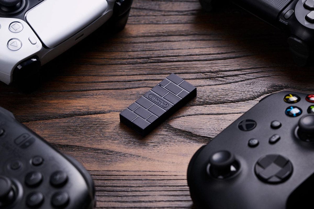 The black 8BitDo USB Adapter 2 sits in the middle of controllers on a wooden table.