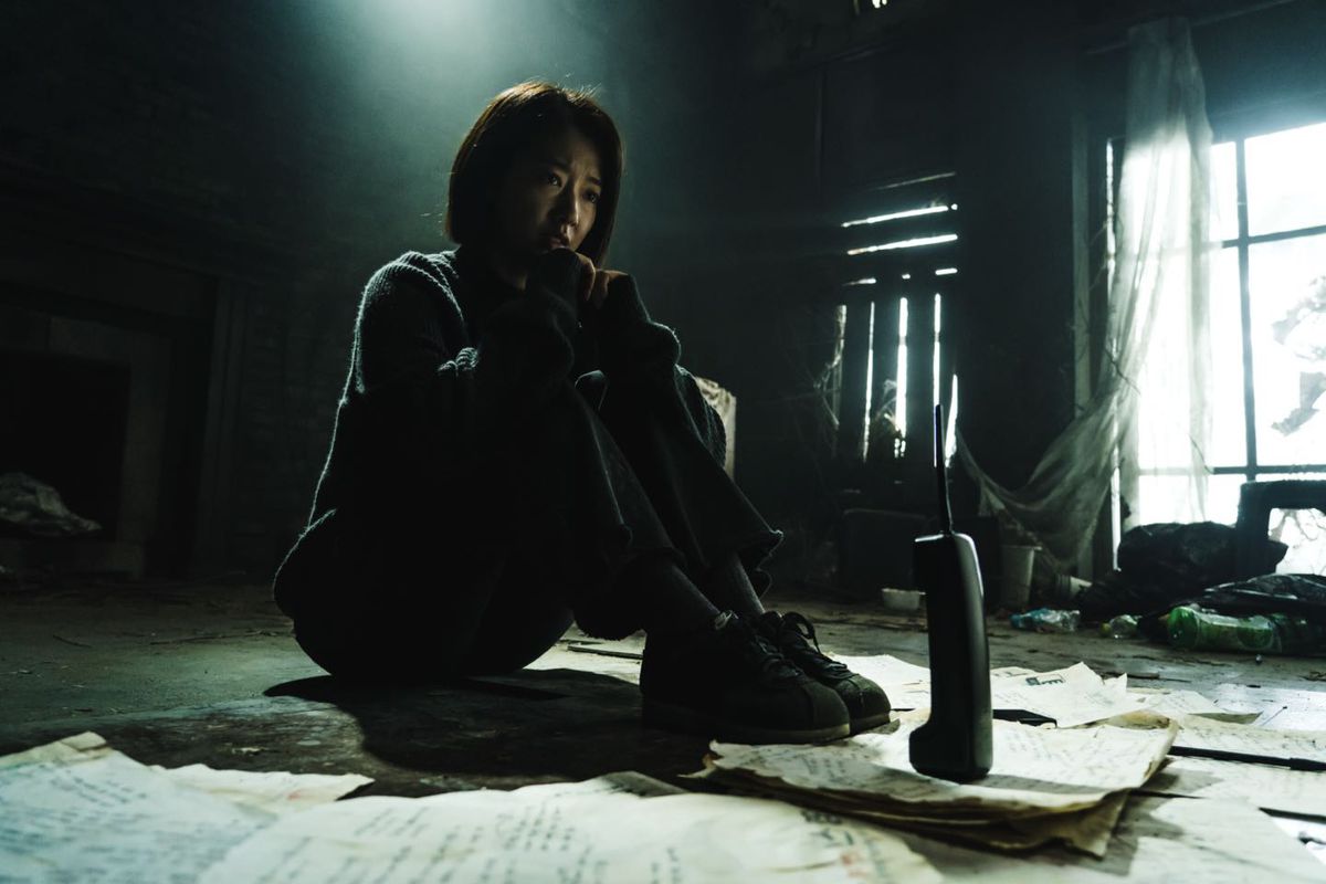 Park Shin-hye sitting across from a cordless home phone in a dark dilapidated living room in The Call (2020)