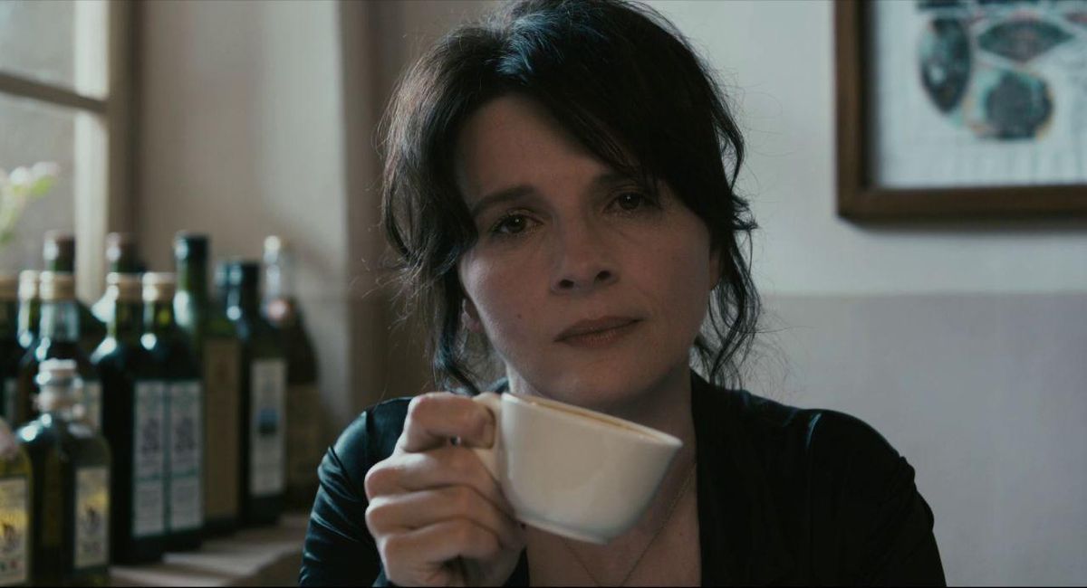 Juliette Binoche looks at the camera and holds a small coffee cup at an angle, tilted down, in Certified Copy.