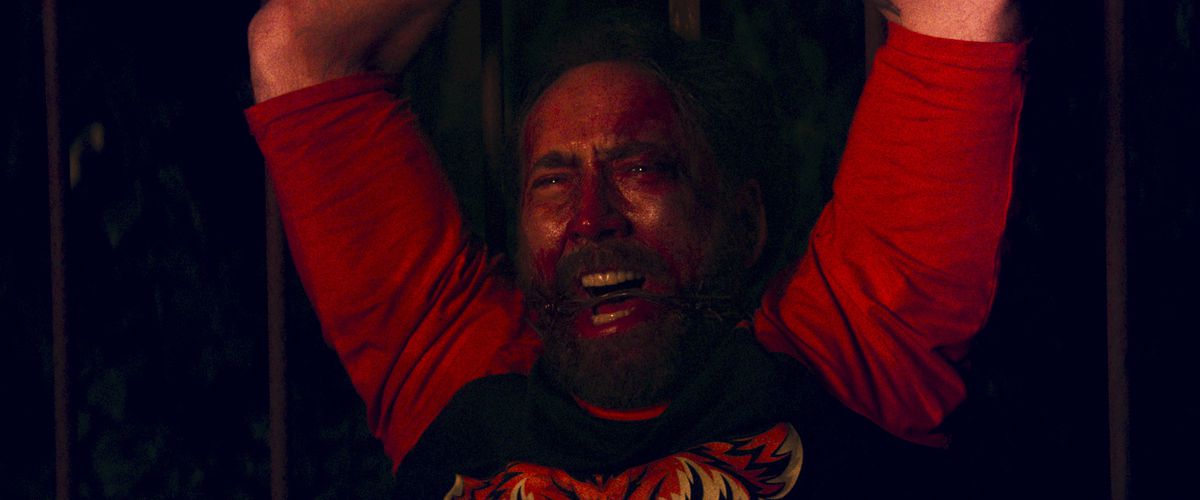 Nicolas Cage as Red Miller, screaming with his hands tied above him while wearing a blood-read shirt with visible bruises and a gag in his mouth in Mandy.