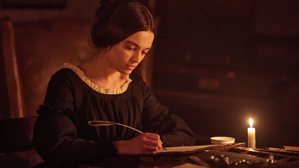 Emma Mackey as Emily Brontë writing on a piece of parchment with a quill pen by candlelight in Emily.
