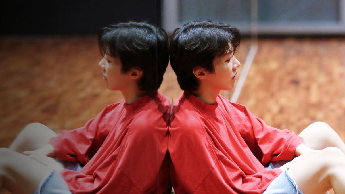 BTS rapper J-Hope leans against a floor-length mirror while wearing jean shorts and a light red long-sleeve shirt. We see his reflection in the mirror.