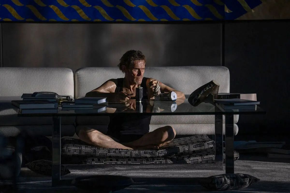 A naked, disheveled man in boxer briefs (Willem Dafoe) sits cross legged in front of a glass table in a darkened living room with light pouring in from a window offscreen in Inside.