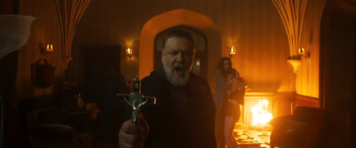 Russell Crowe holds up a cross with flames behind him in The Pope’s Exorcist.