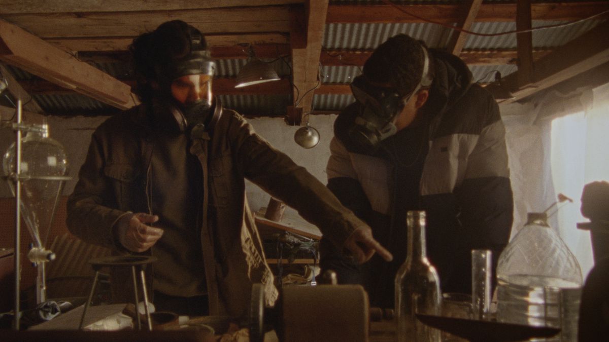 Two people wearing gas masks work with chemicals, while one points, in How to Blow Up a Pipeline.