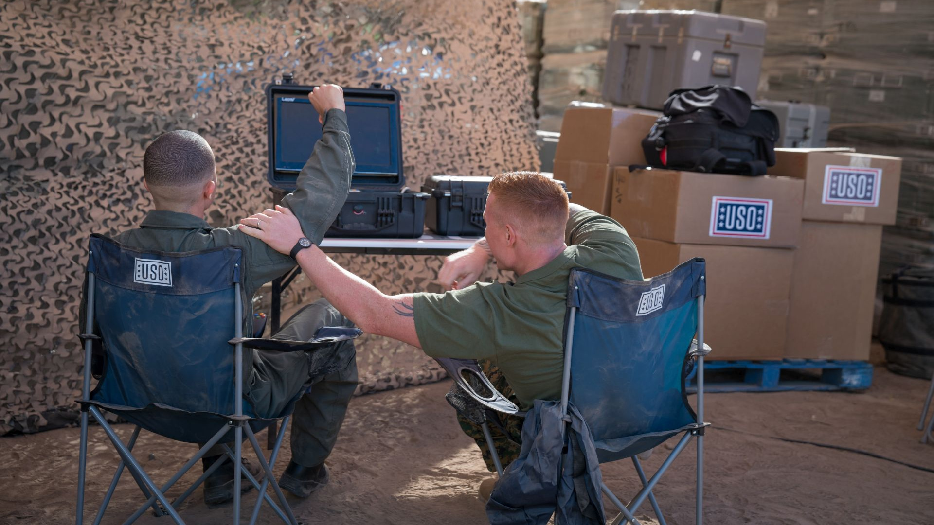 Two service members sitting in camping chairs celebrate at a military encampment where they are able to play video games using mobile gaming kits.