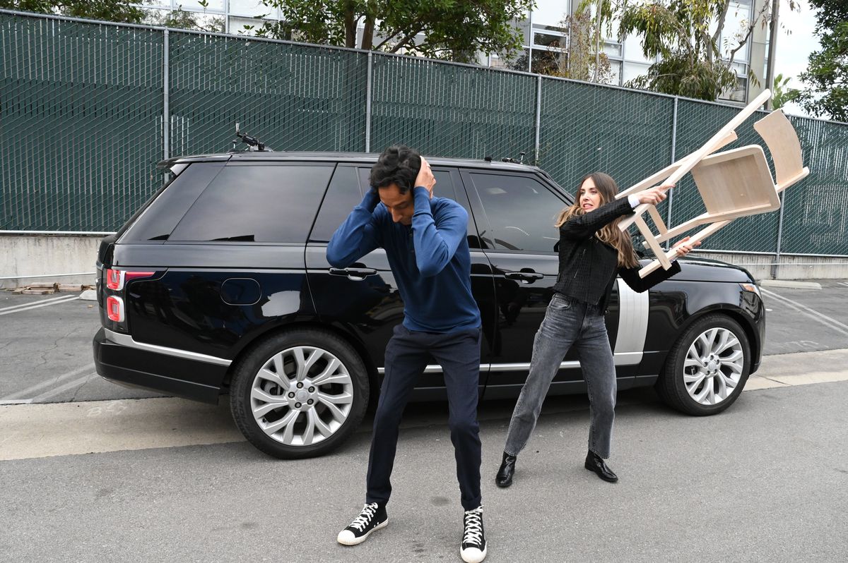 Danny Pudi holding his ears and crouching a bit while Alison Brie comes up behind him with a chair about to hit him. The Carpool Karaoke car is behind them. 