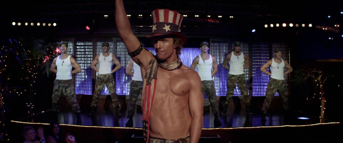 A shirtless Matthew McConaughey in a red, white, and blue striped hat with stars and handkerchief stands in front of a stage of men in white shirts and camouflage pattern pants standing at attention in Magic Mike.