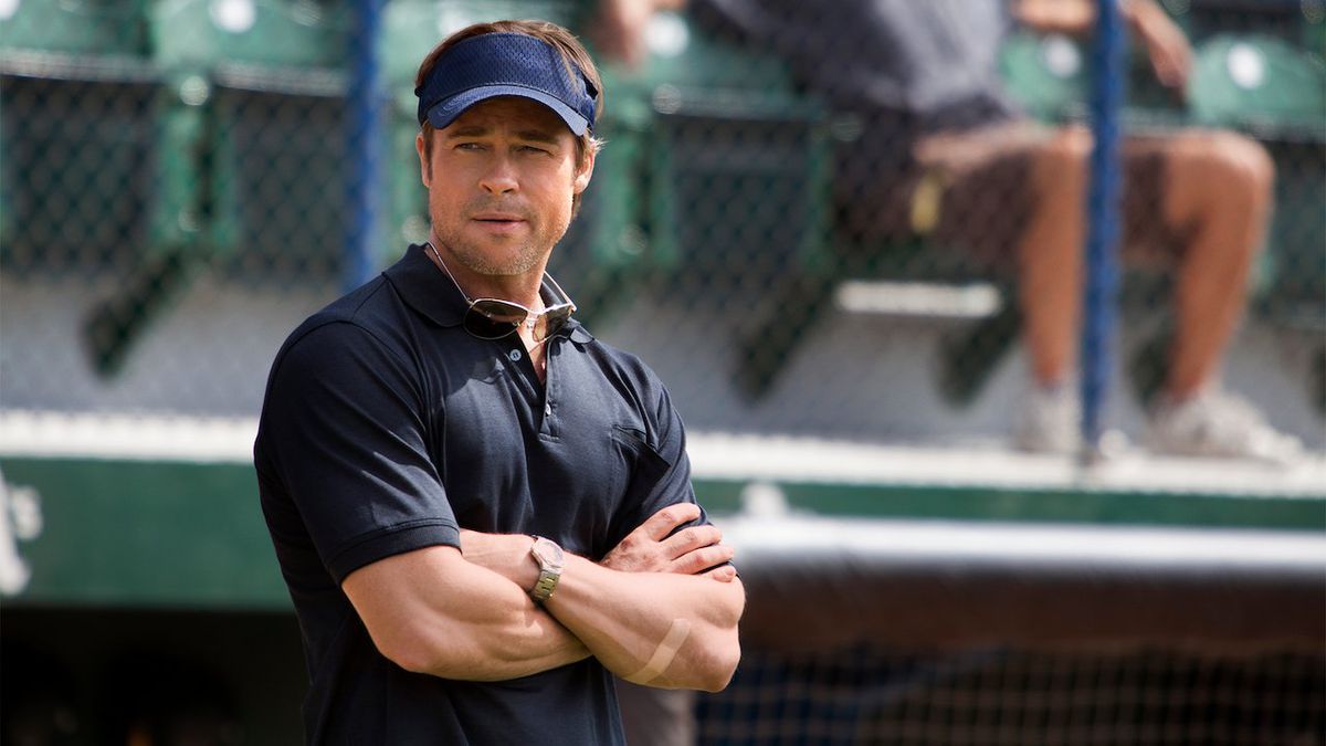 A man in a navy blue polo (Brad Pitt) wearing a blue hat while standing on the side of a baseball field.