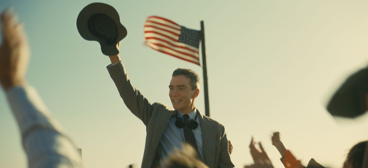 J. Robert Oppenheimer (Cillian Murphy) grins and waves his hat at a cheering outdoor crowd while standing under an American flag in Christopher Nolan’s Oppenheimer