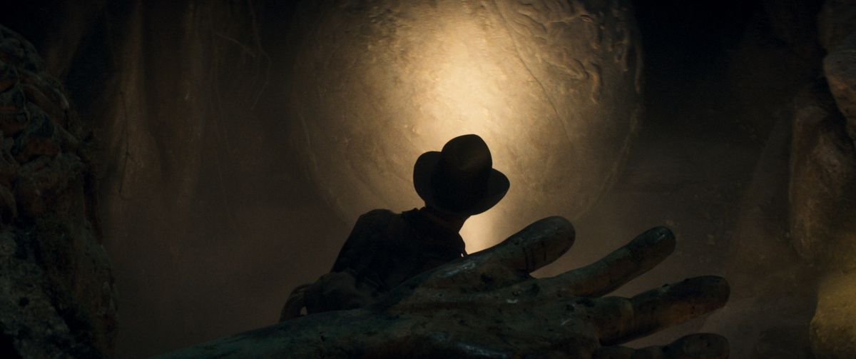 Silhouette of Indiana Jones exploring a tomb, the hand of a corpse is in the foreground, also shadowed.