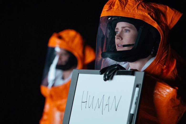 Amy Adams wearing an orange hazmat suit and holding a dry erase board with the word “human” written on it in Arrival