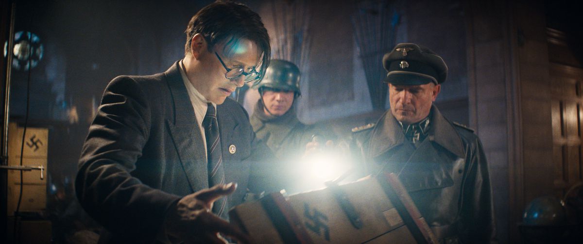 Mads Mikkelsen as Doctor Jürgen Voller opens a crate while two Nazi soldier overlook, shining a flashlight inside in Indiana Jones and the Dial of Destiny.